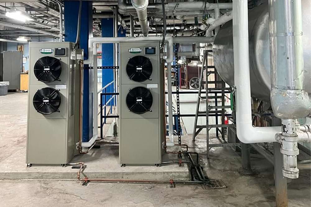 Two air conditioning units in an industrial setting, next to piping and a large metal tank.