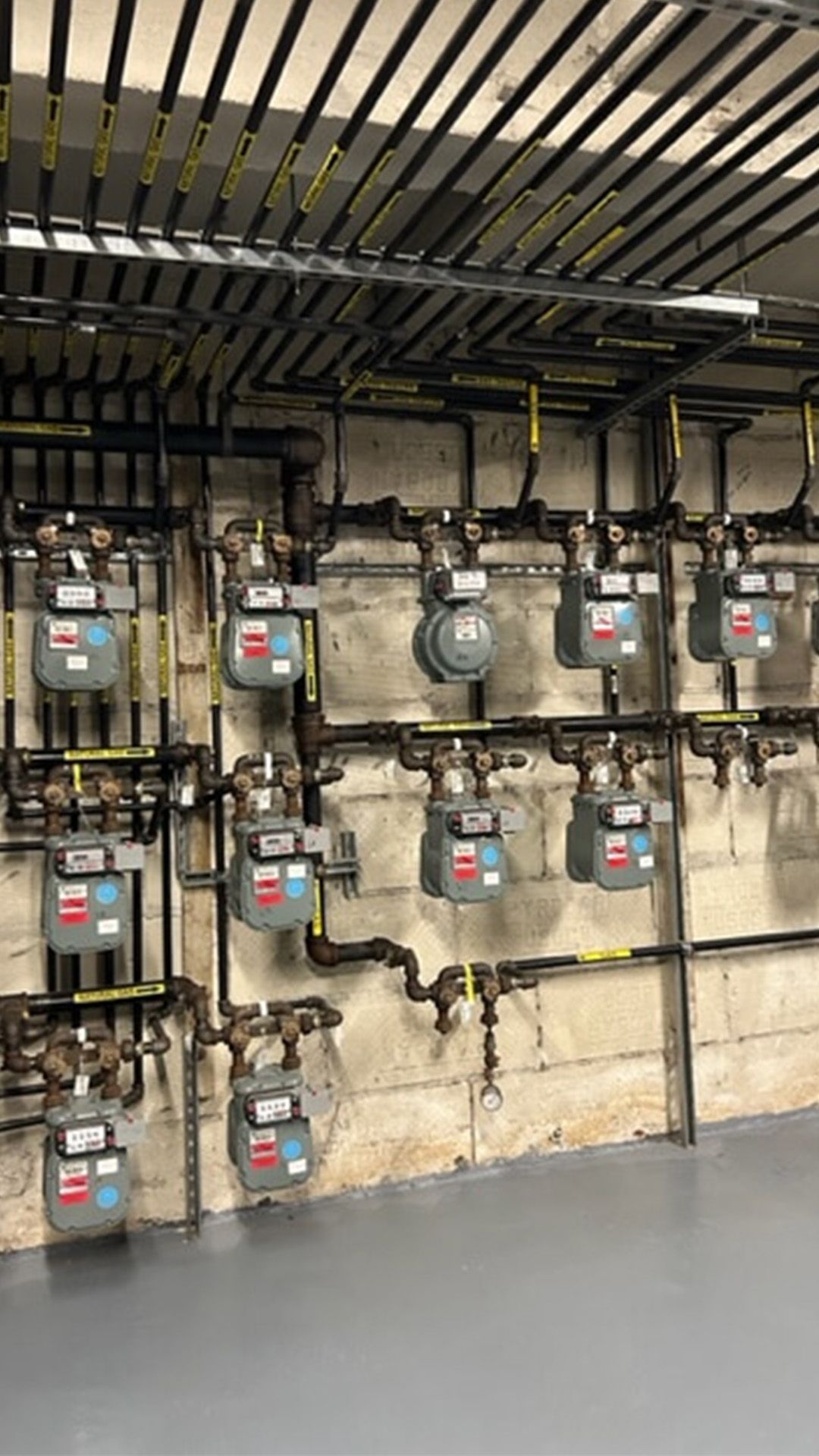Gas meters lined up on a concrete wall, with pipes and valves overhead.
