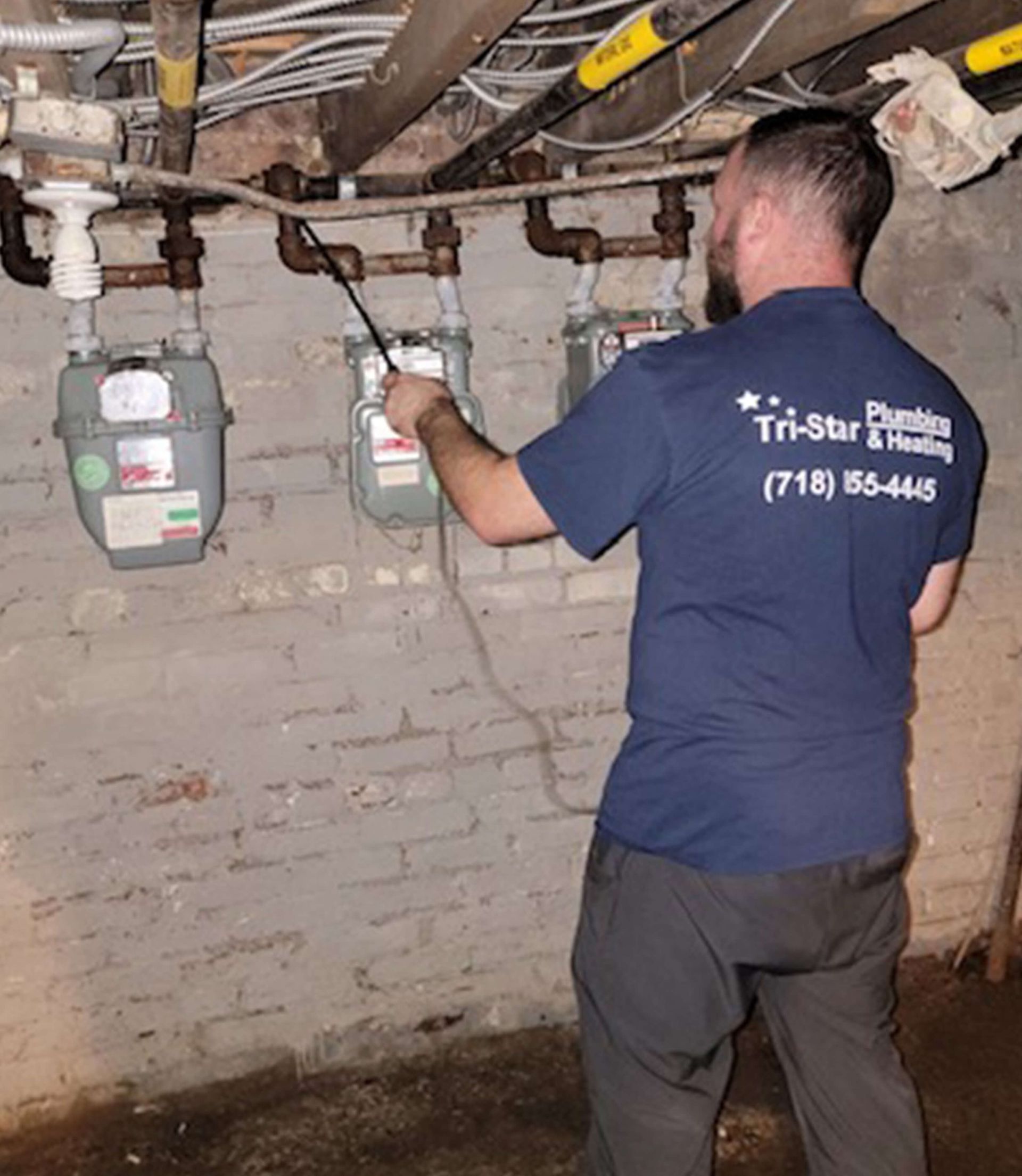 Plumber inspecting gas meters in a basement; wearing a Tri-Star Plumbing shirt and pointing a wand.