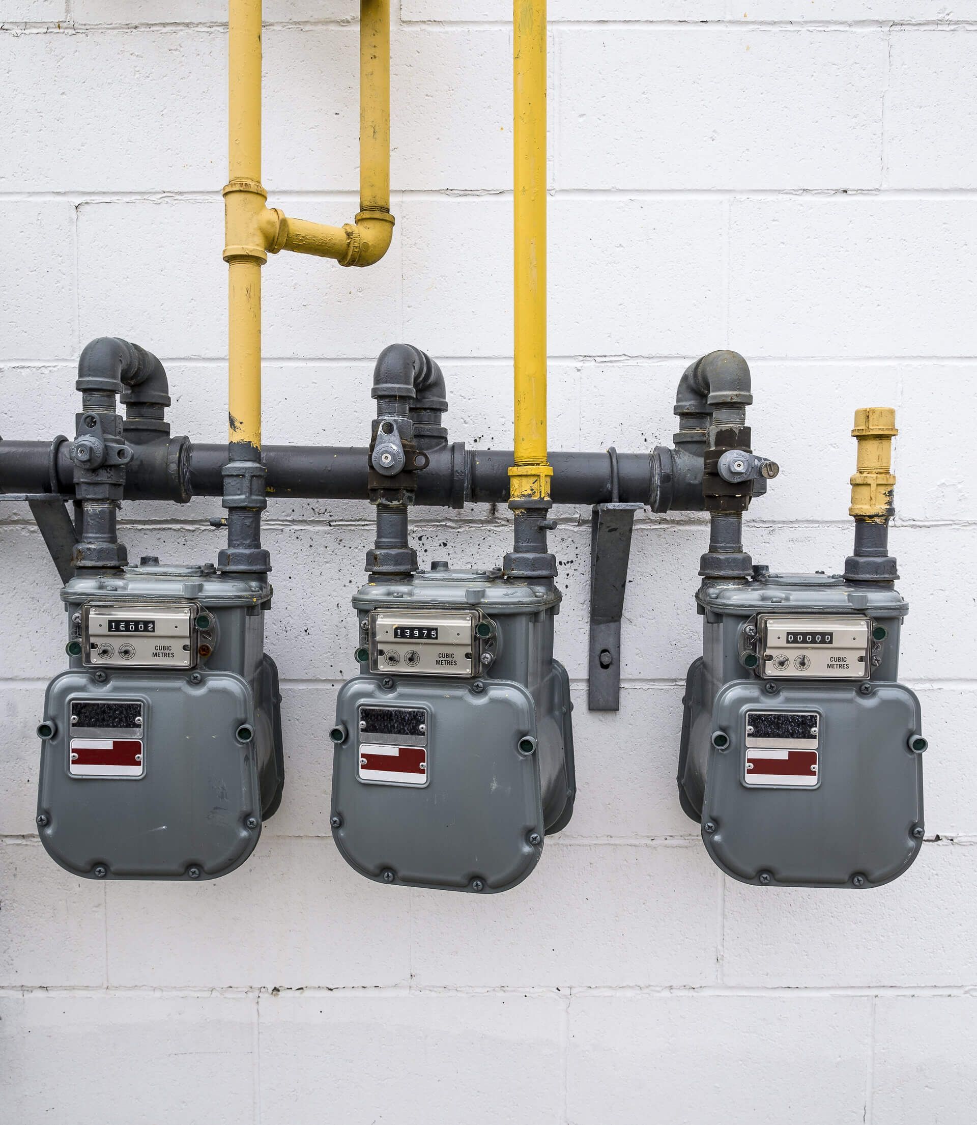 Three gray gas meters mounted on a white brick wall with yellow pipes.