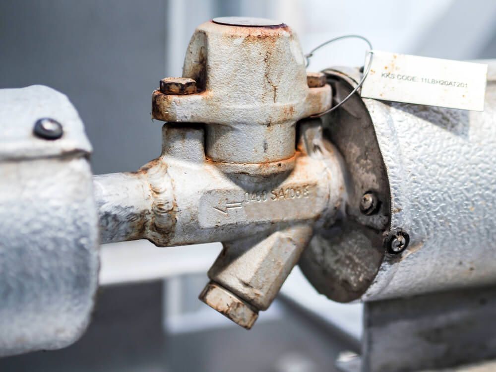 Close-up of a rusty, white industrial valve connected to insulated pipes, with a hanging tag.