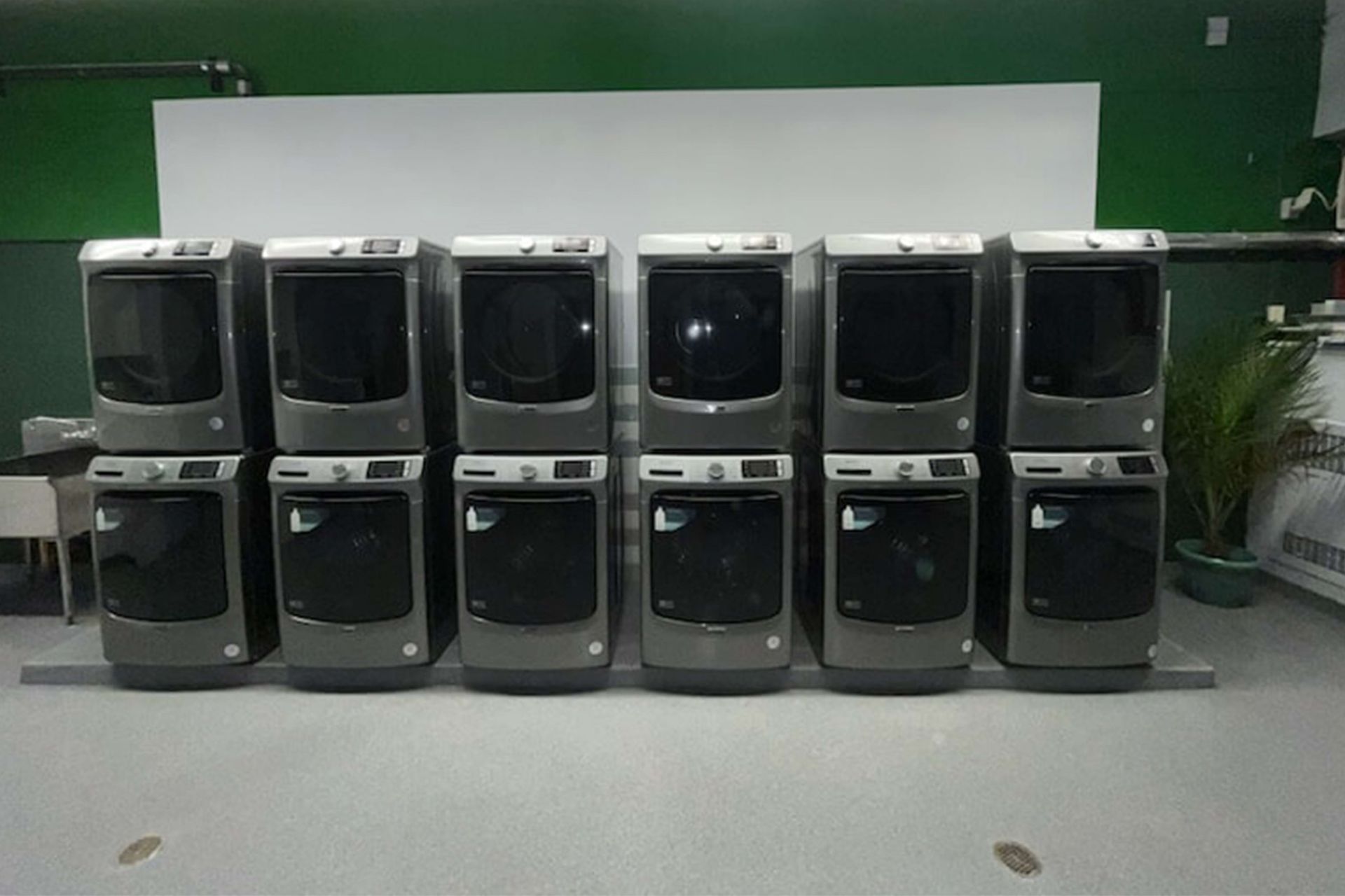 Row of stacked washing machines in a commercial laundry setting; gray machines against a white wall.