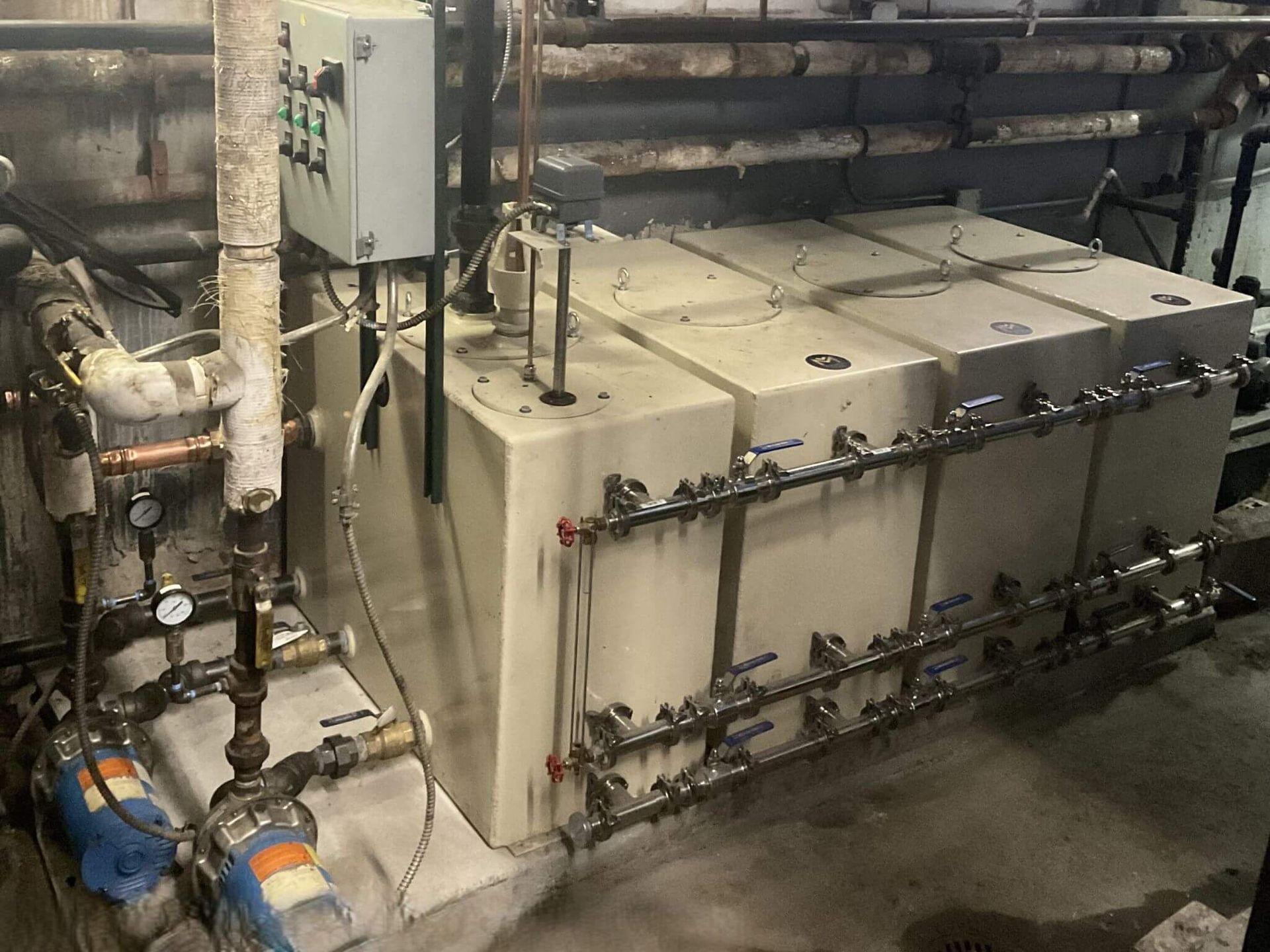 Industrial tanks and piping in a utility room. Tanks are beige, pipes are metallic, and the setting appears utilitarian.
