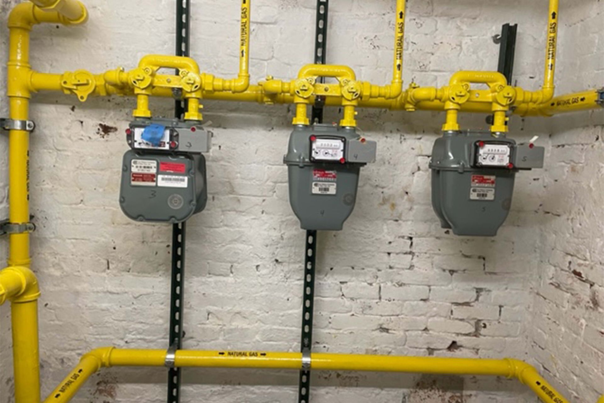 Three gray gas meters mounted on a brick wall with yellow pipes.