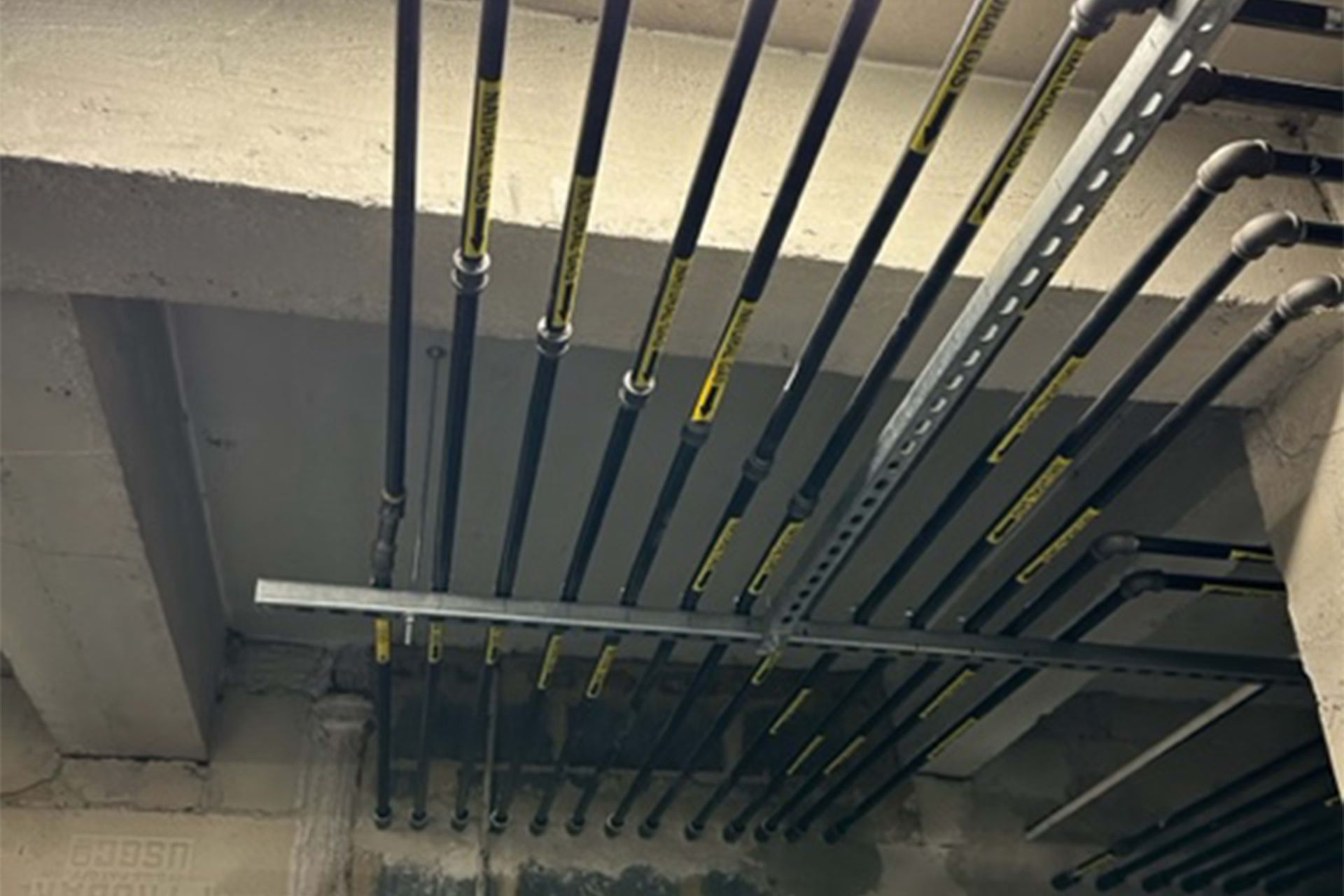 Black pipes with yellow labels are mounted on a metal bracket beneath a concrete ceiling.