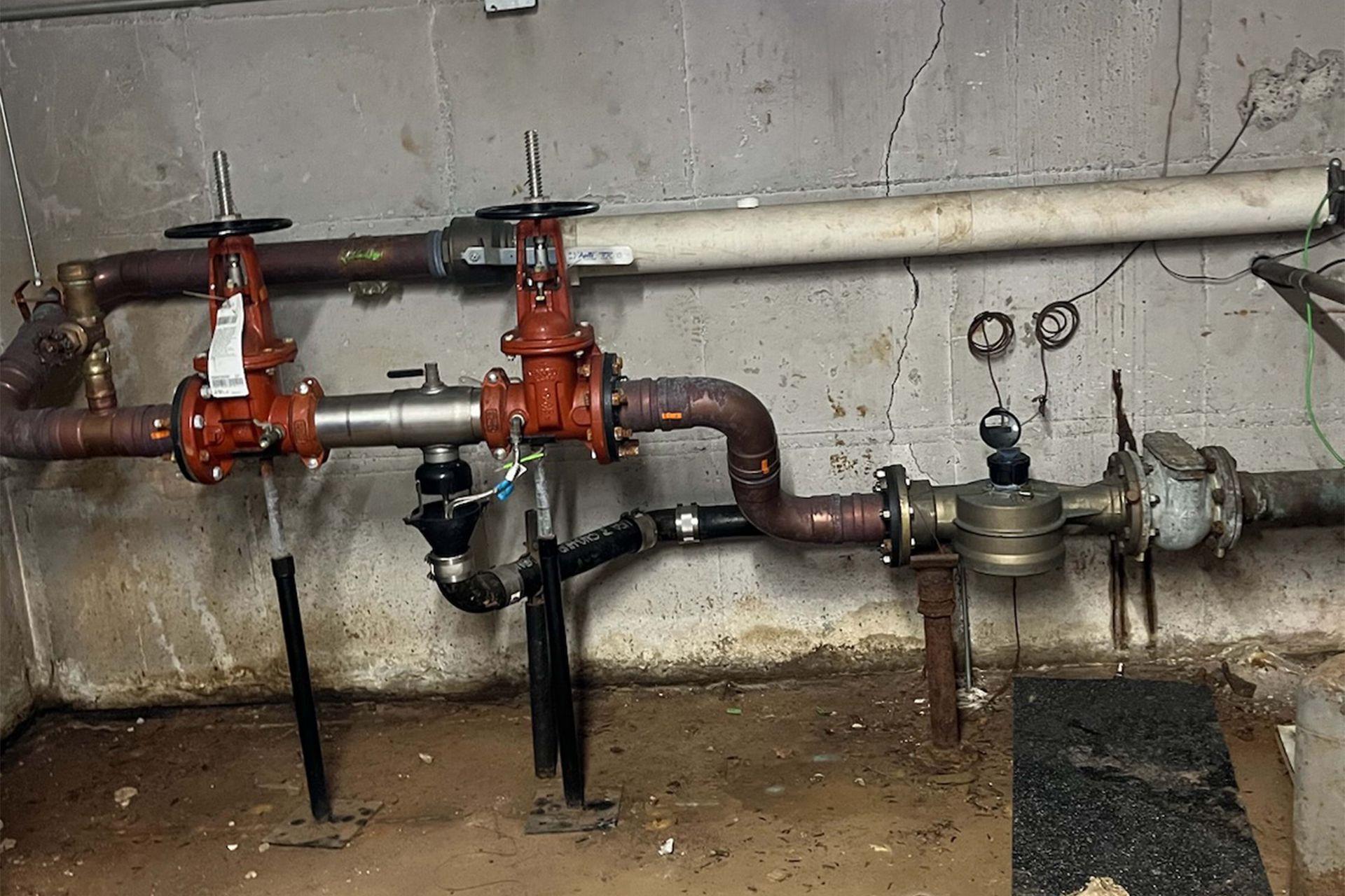 Pipes and valves in a basement setting. Two red valves are visible, along with black and brown pipes against a concrete wall.