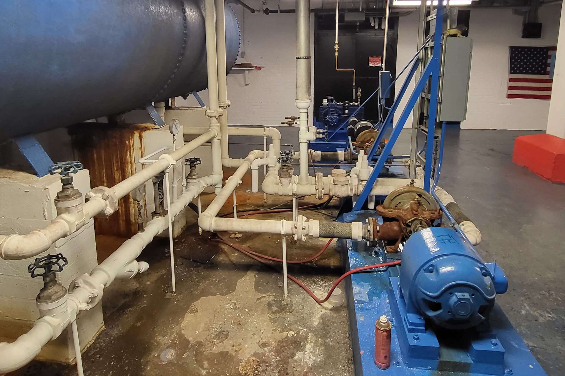 Blue machinery and pipes in a room. Rusty metal and wet floor are visible. An American flag hangs on the wall.