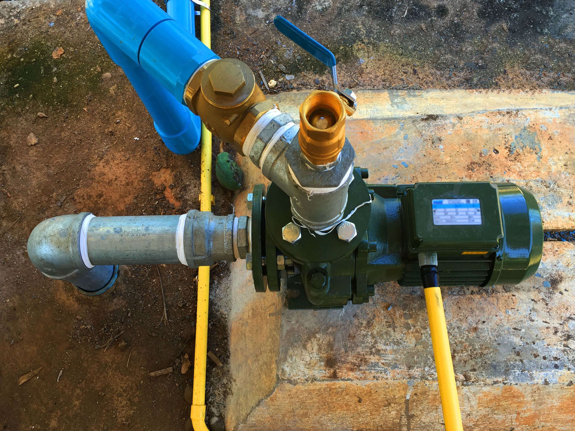 Water pump with blue and brass pipes, a yellow hose, and a green motor, outdoors.