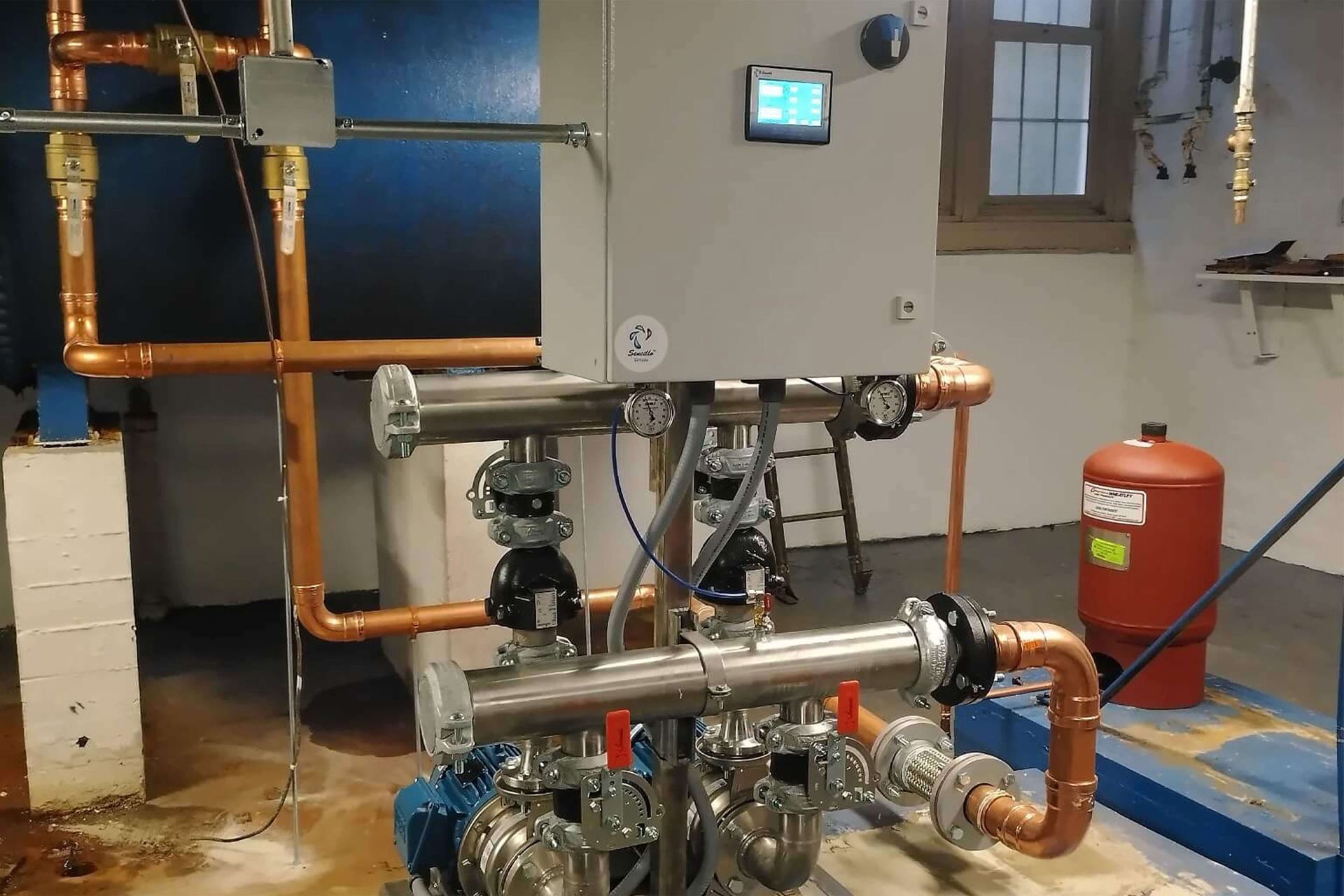 Complex plumbing system with copper pipes, stainless steel components, control box, and a red expansion tank.