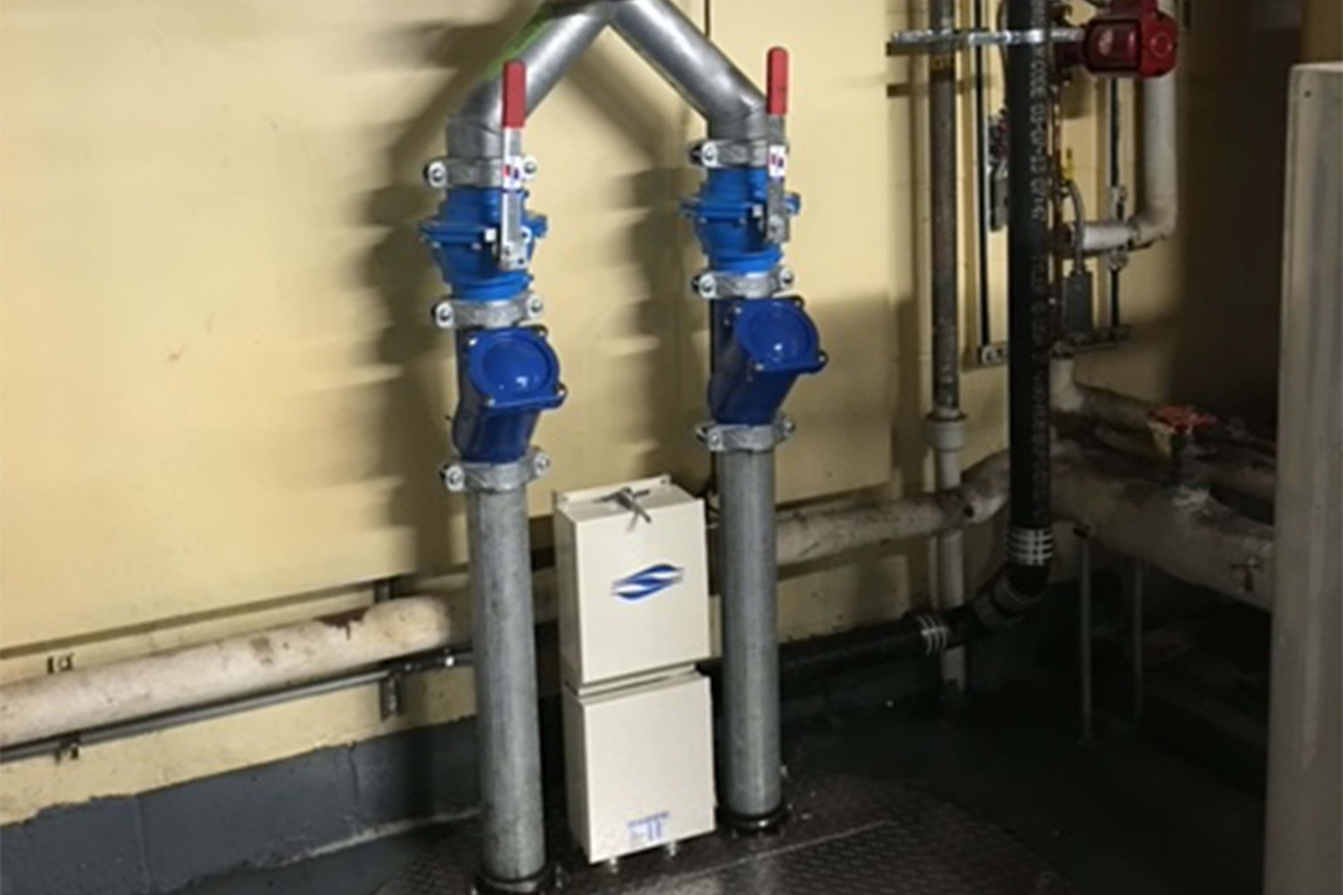 Blue valves on vertical pipes connected to overhead piping in a room with a metal floor.