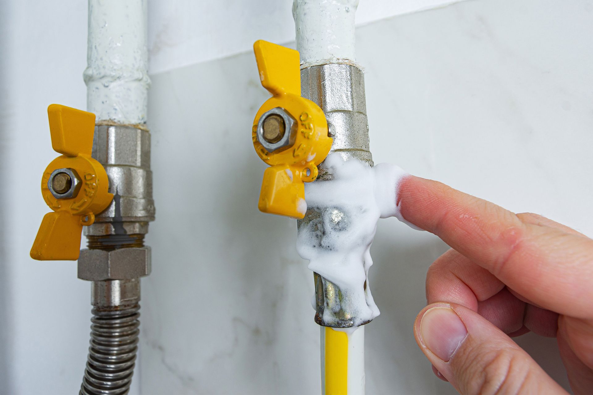 A hand touching a soapy white foam on a gas pipe joint; yellow valves and pipes are visible.