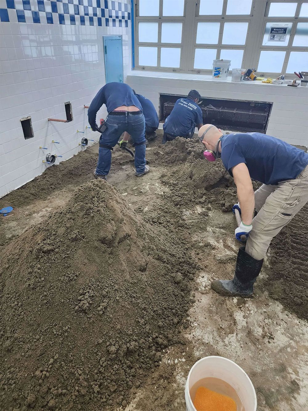 Workers in a pool, spreading material on the floor. White tiles on the wall, blue trim, windows in background.