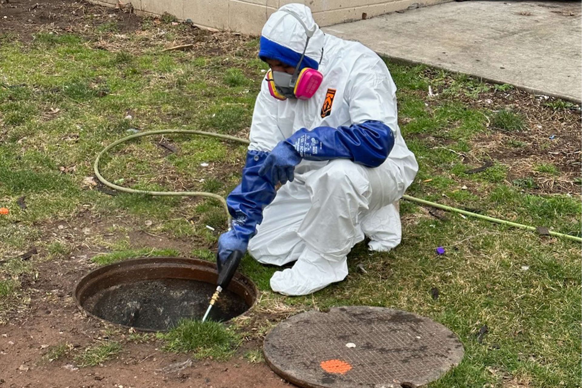 Person in protective suit examining an open manhole in grassy area.