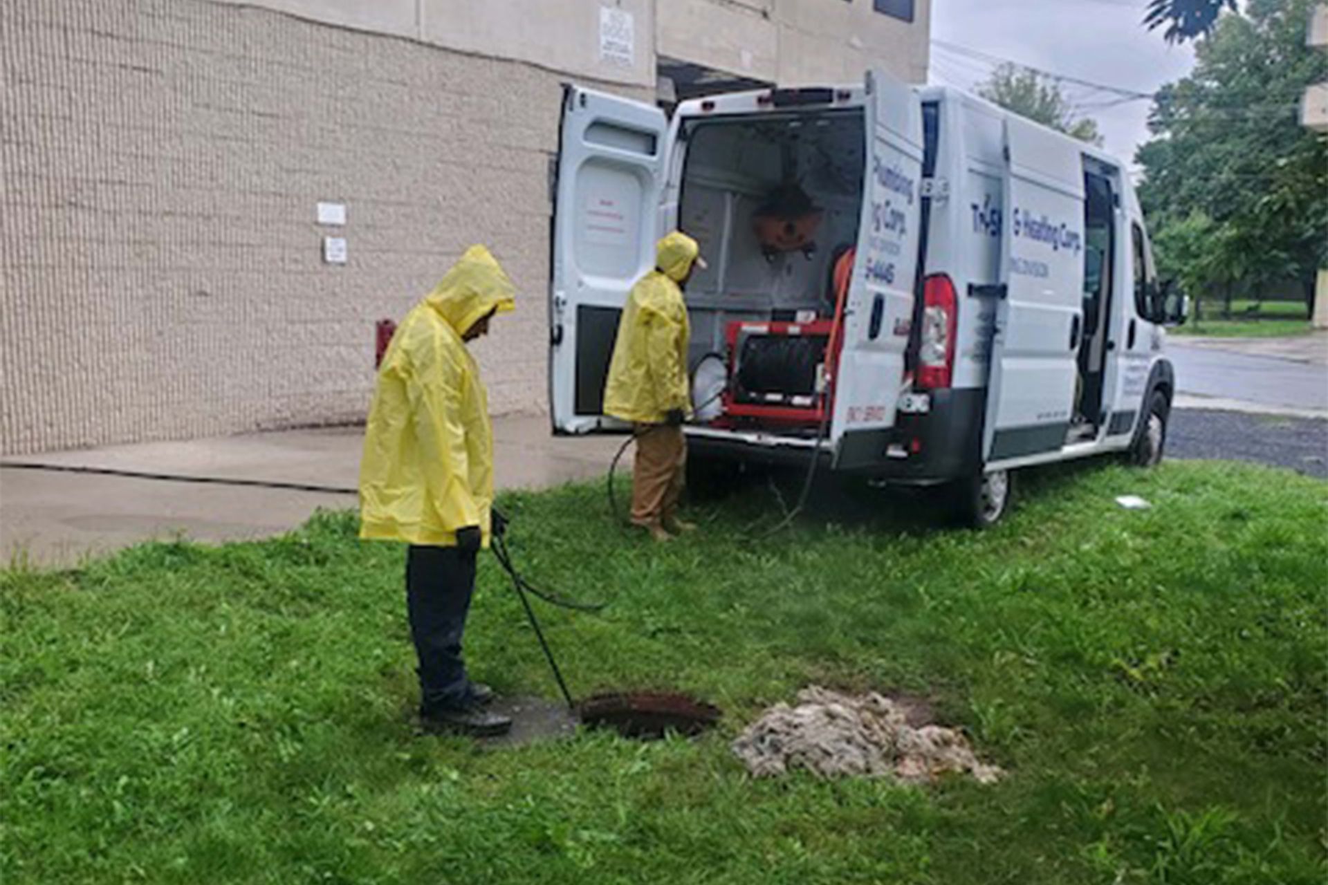 Two people in yellow raincoats working on a drain near a white van marked 