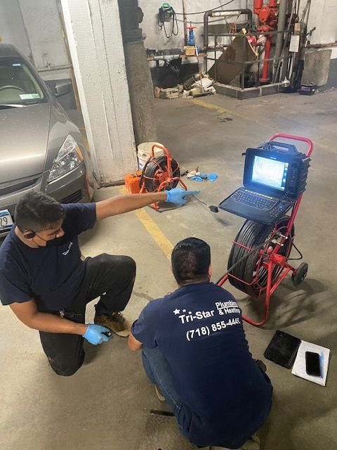Two plumbers inspect sewer line with camera in a garage. One points, the other monitors a screen.