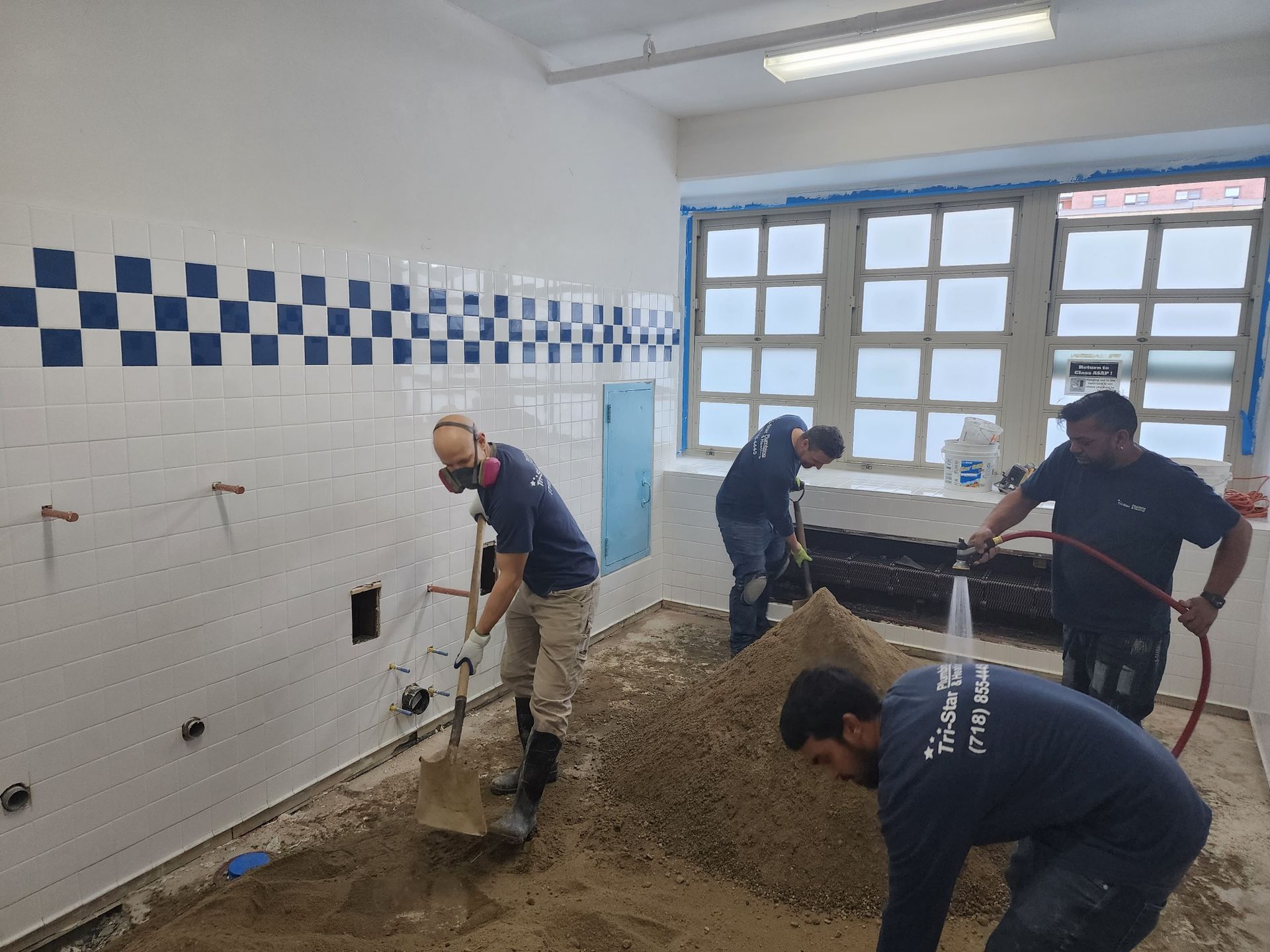Four workers in a room, removing sand with tools. White and blue tiled walls, window, water hose.