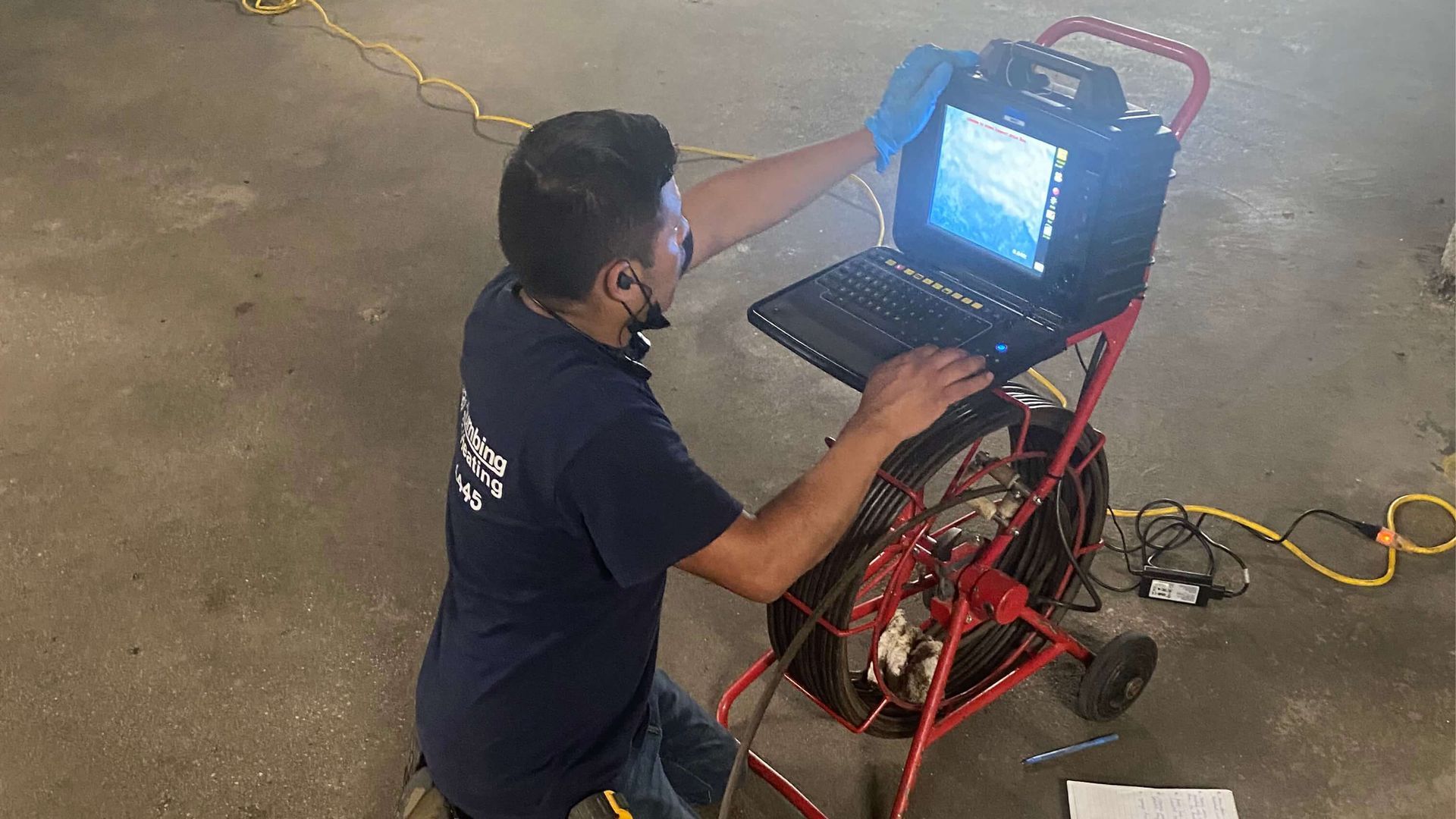 Man using a laptop connected to a sewer camera cart on a concrete floor.