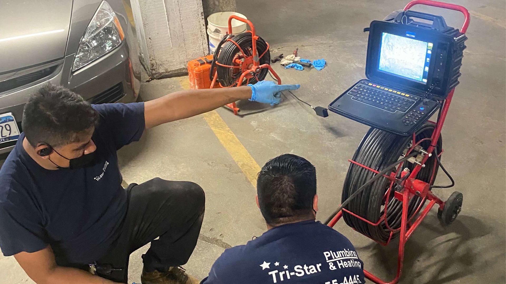 Two plumbers using a sewer camera in a parking garage. One points at screen; the other looks on.