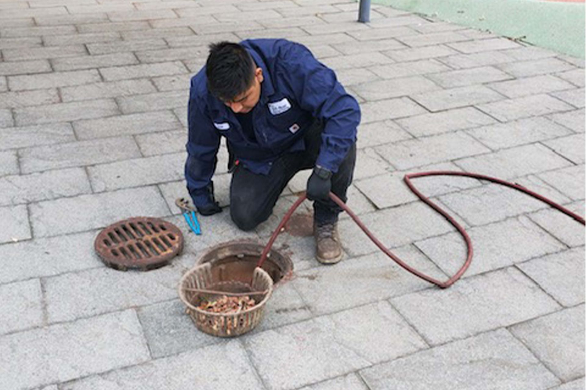 Man kneeling, cleaning a sewer drain on a brick sidewalk. Brown hose and a basket of debris are visible.