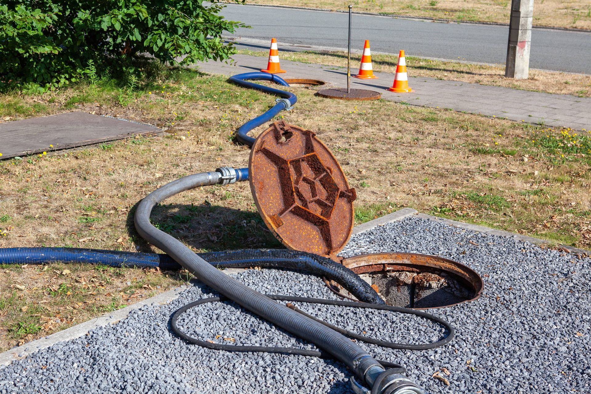Sewer work: Open manhole with hoses, rusty cover, orange cones on a gravel area next to a road.