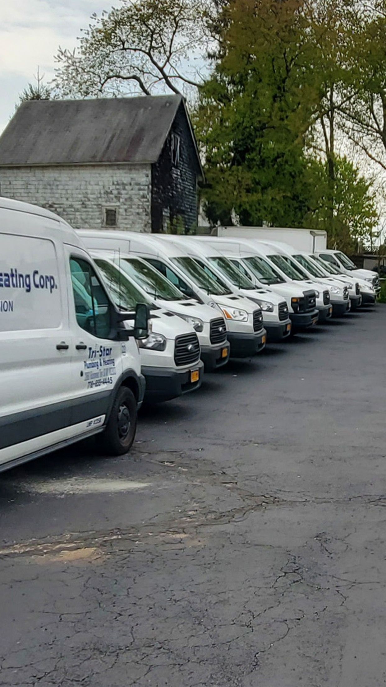Line of white work vans parked on asphalt with a gray building and trees in the background.