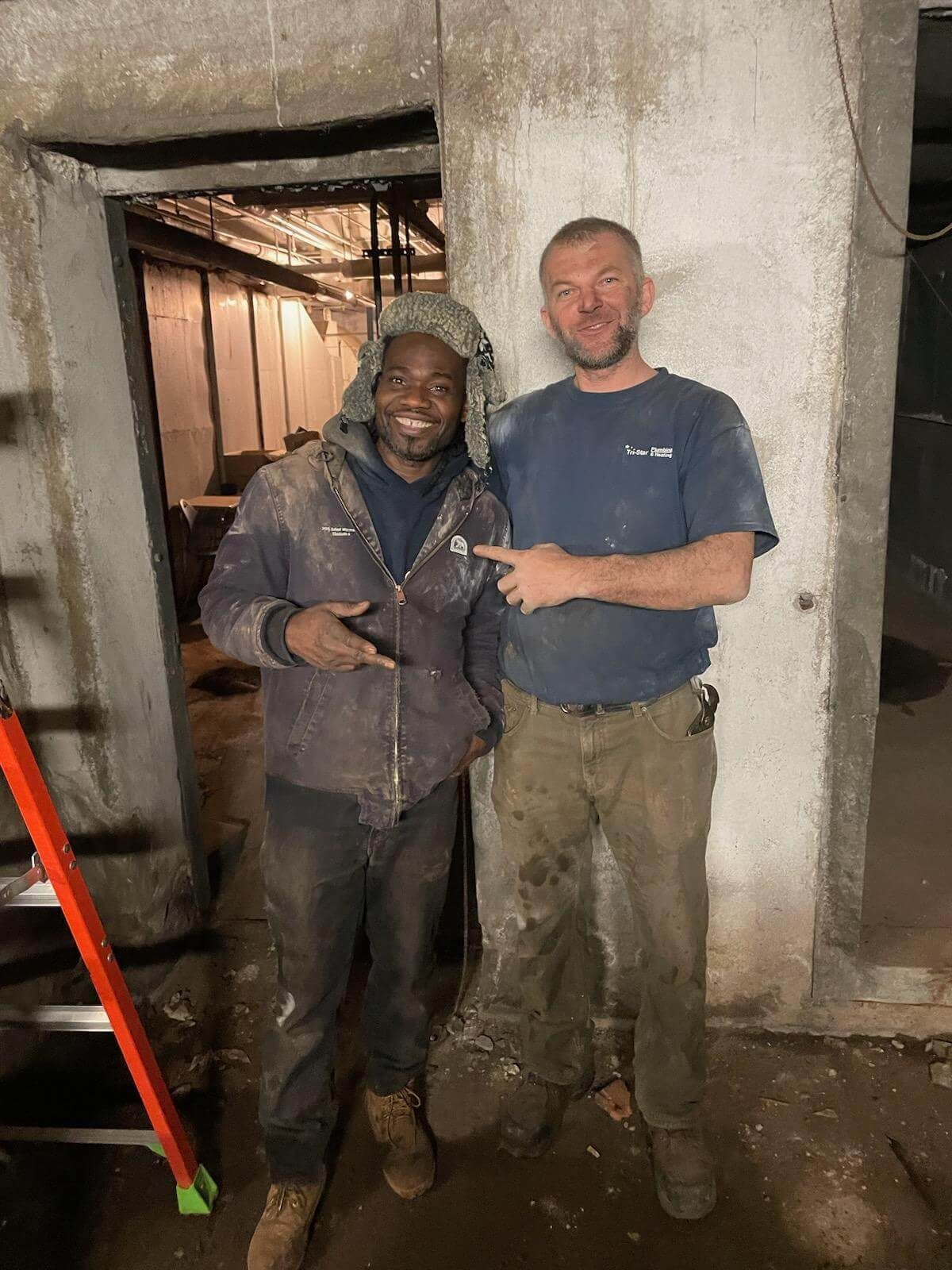 Two men in work clothes pose indoors. One points; the other smiles, both in a grimy, concrete environment.