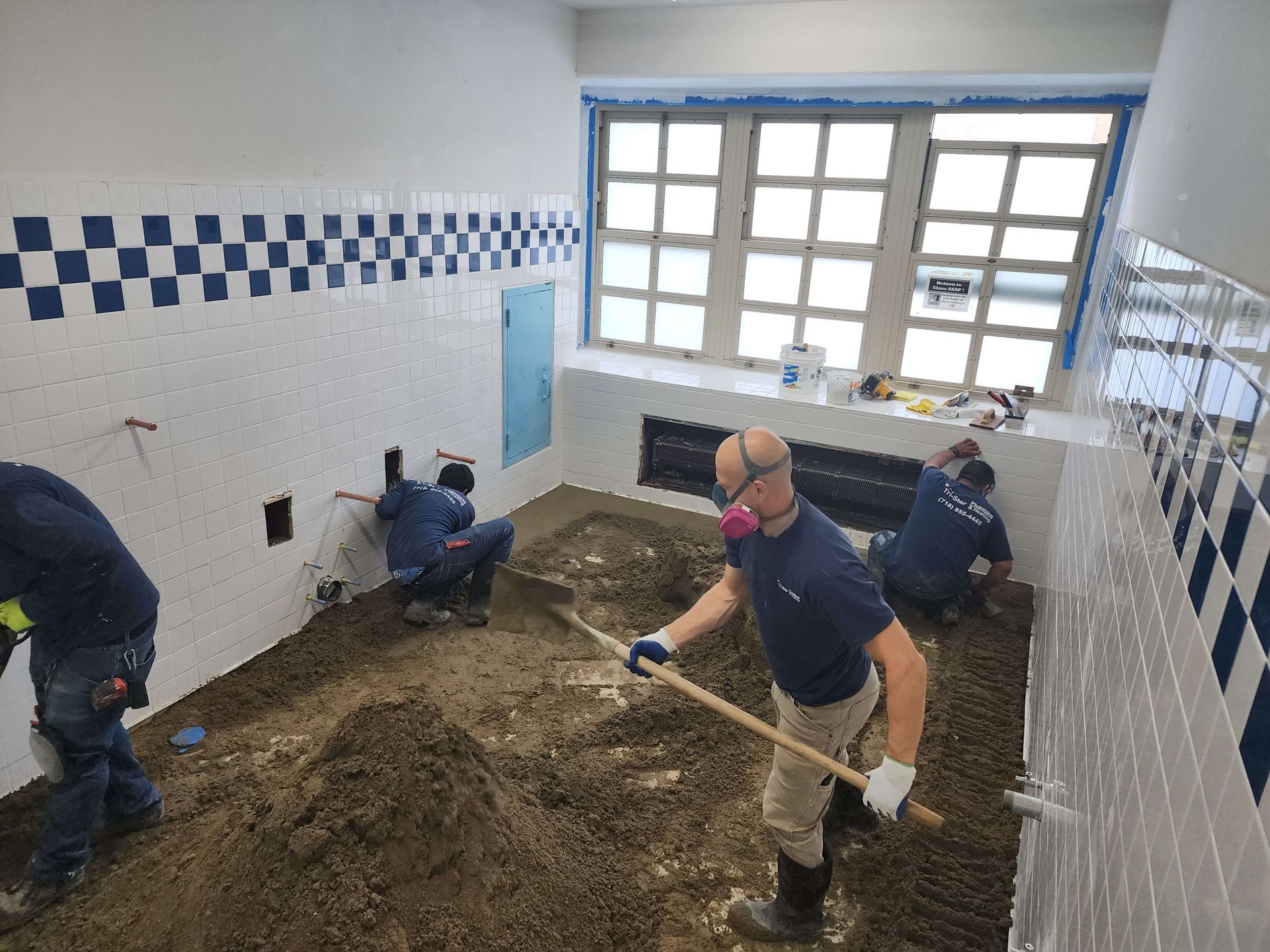Workers inside a bathroom, shoveling dirt. White and blue tile walls. Window in the background.