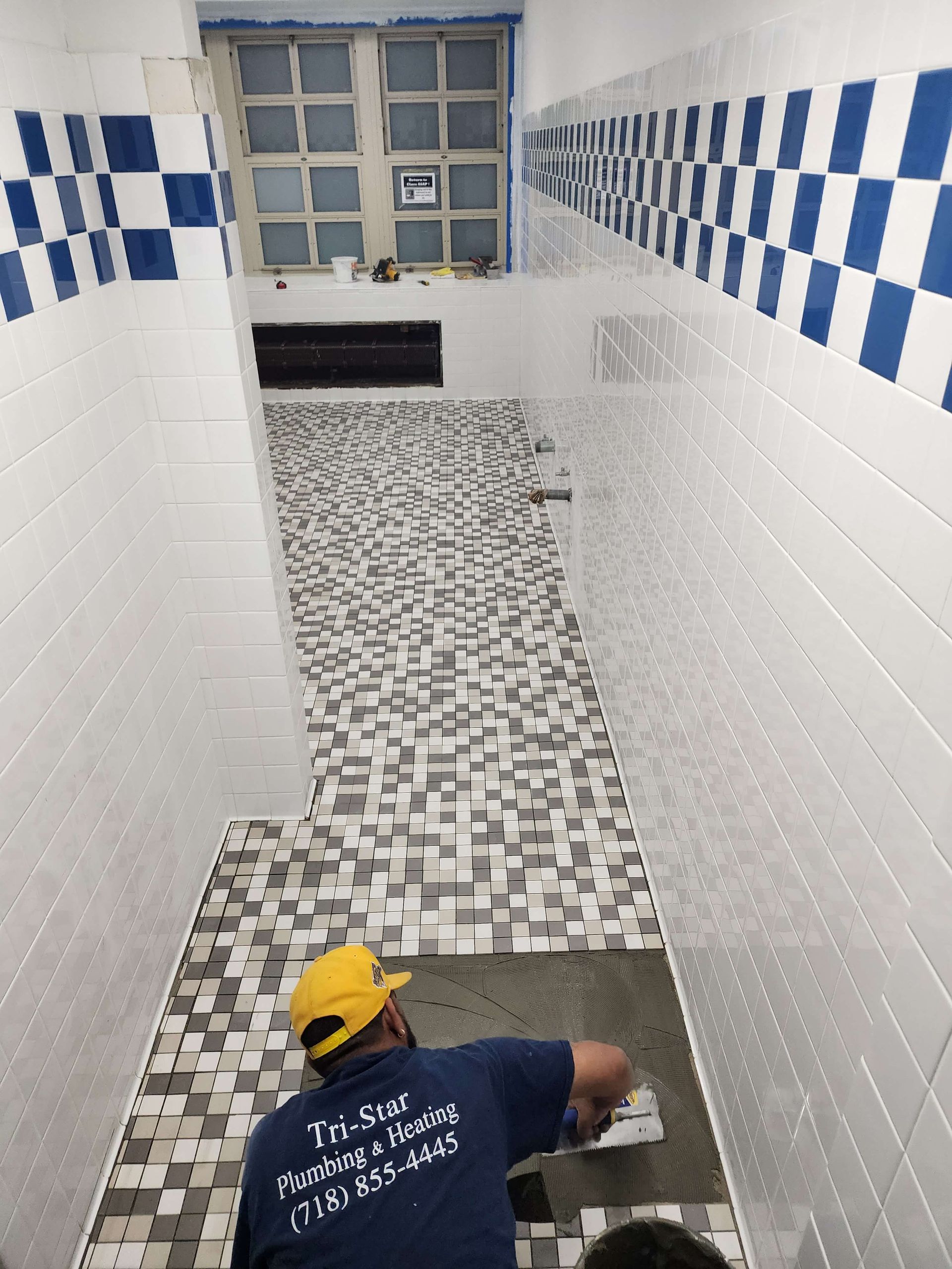 Person tiling a bathroom floor with multi-colored tiles. Blue and white tiled walls.