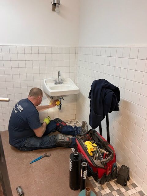 Plumber working on a sink in a tiled bathroom. He kneels on the floor, surrounded by tools.