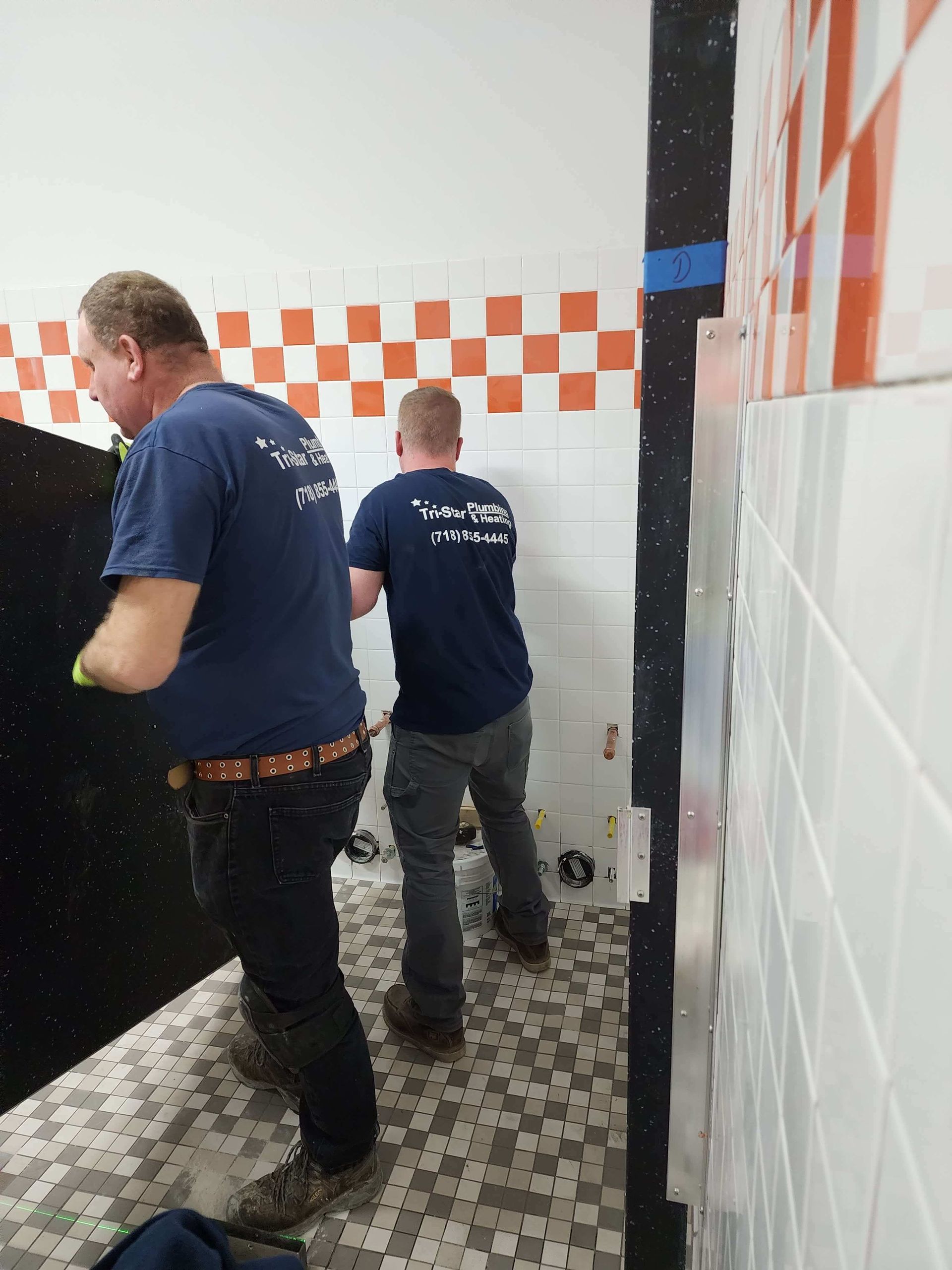 Two men installing wall tiles in a bathroom, one holding a black object, checkerboard design above white tiles.