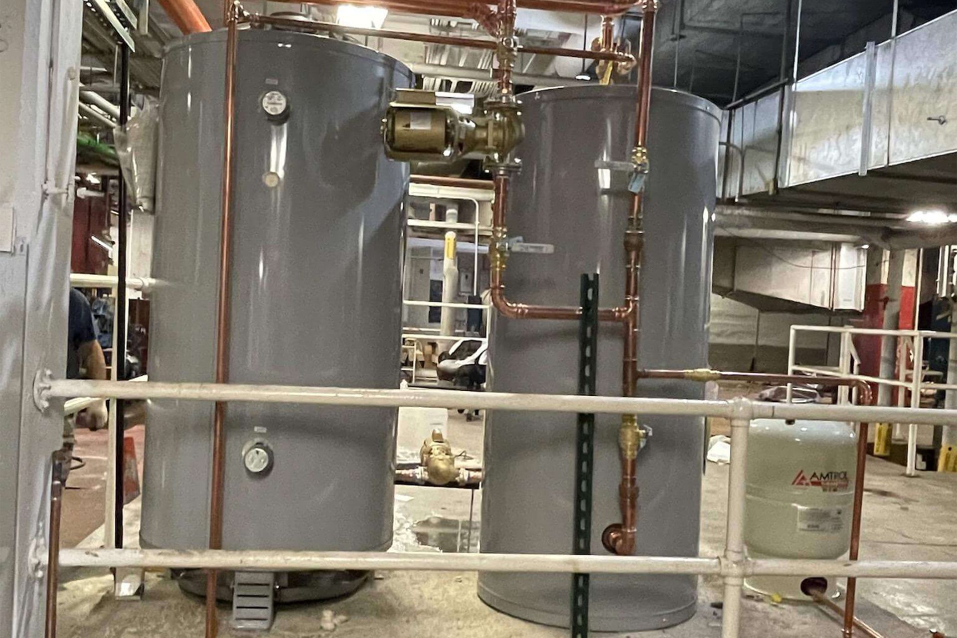 Two large gray water heaters with copper pipes, gauges, and a small expansion tank, indoors.