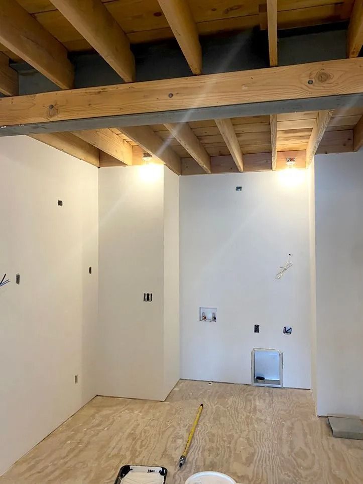 Interior of a room under construction with white walls, exposed wooden beams, and plywood flooring.