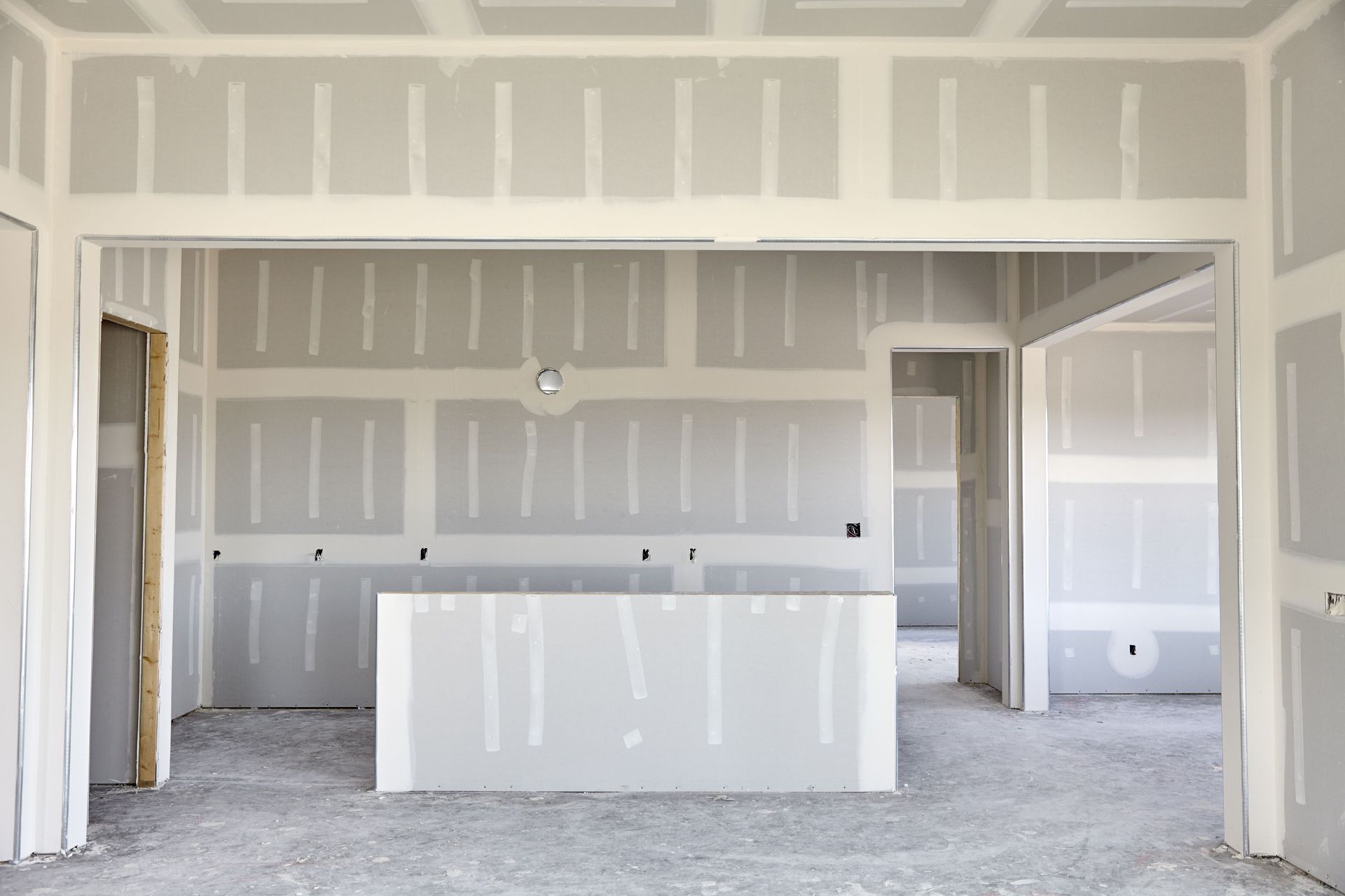 Room under construction with drywall walls and ceiling, two windows, and exposed light fixtures.