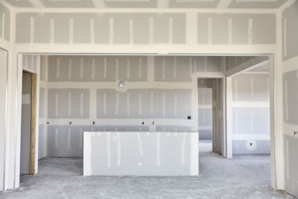 Room under construction with drywall walls and ceiling, two windows, and exposed light fixtures.