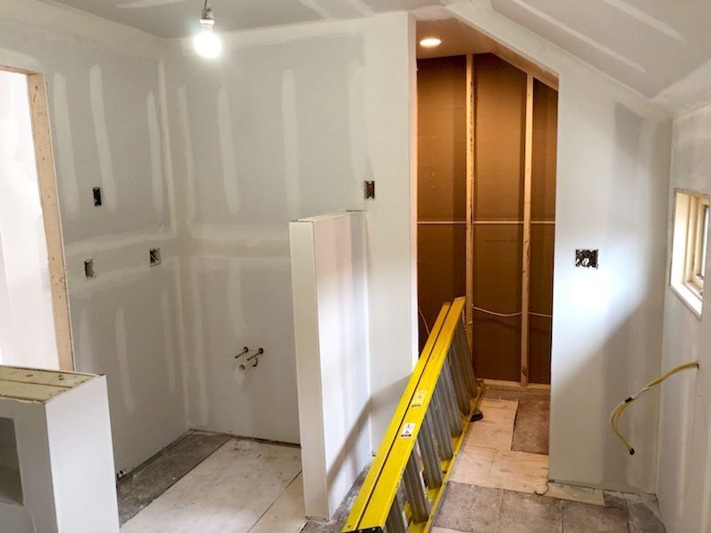 Interior room under construction, drywall installed. A yellow ladder leans against a wall leading to a doorway.