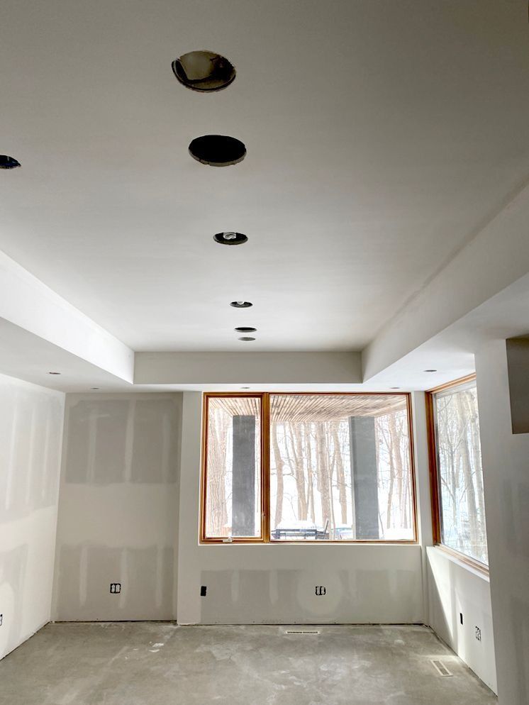 Interior of a room under construction with recessed ceiling lights and large windows covered in plastic.