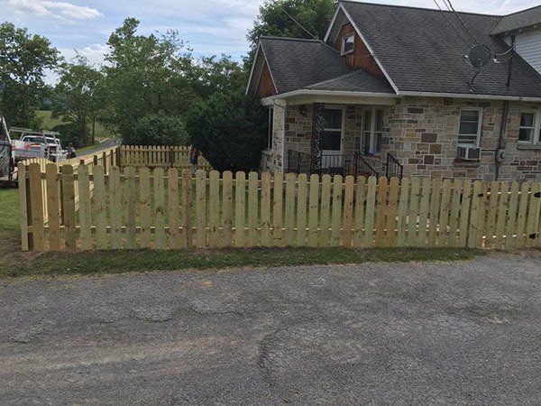 House with stone facade behind a pale wooden picket fence by a road and trees