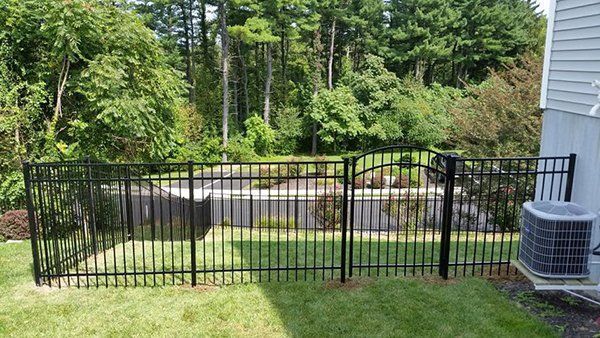 Black metal fence around a lawn and garden beside a house, with trees and shrubs in the background