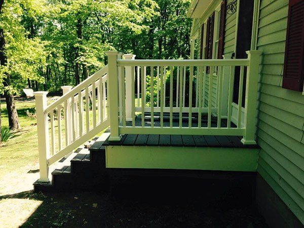 White porch railing and steps beside a green house, with a wooded yard in the background.