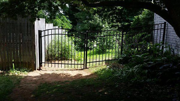 Open black metal gate at the end of a sunlit garden path surrounded by trees and shrubs