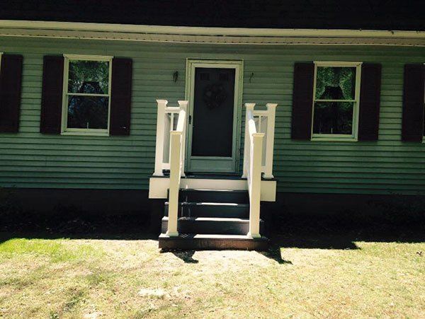 Front of a green house with a small porch, steps, and two windows with dark shutters
