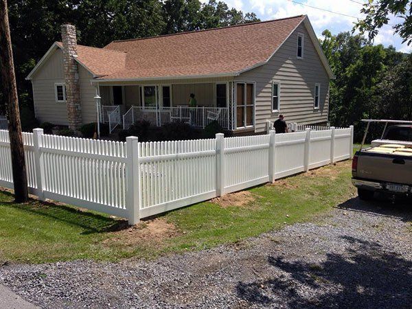 White picket fence around a beige house with a red roof and driveway on a sunny day