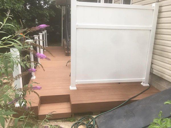 Wooden deck beside white fence and house siding, with purple flowers and a black hose on the ground.