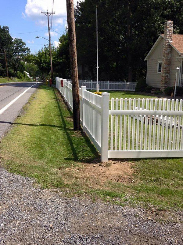 White picket fence beside a rural road with a beige house and green lawn under a bright sky