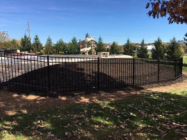 Black metal fence along a paved area in a sunny park with trees and a blue sky