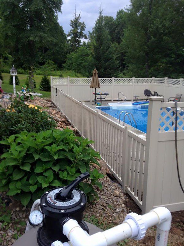 Backyard pool behind a white fence with patio umbrellas and green shrubs beside a pool pump and pipes