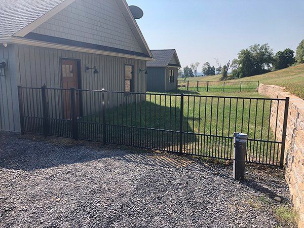 Black metal gate beside a beige house, opening to a gravel driveway and grassy field.