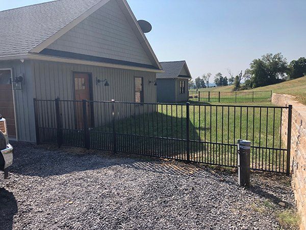 Residential gate beside a beige house, opening to a grassy yard and gravel driveway.