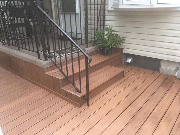 Wooden front porch steps with black metal railing beside tan house siding and a small potted plant.