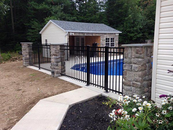 Backyard pool enclosed by black metal fence beside stone pillars and a paved walkway